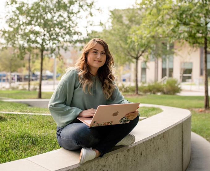 A CU Denver student sitting outside on a curved concrete bench with a laptop in her lap, smiling and surrounded by trees on campus.