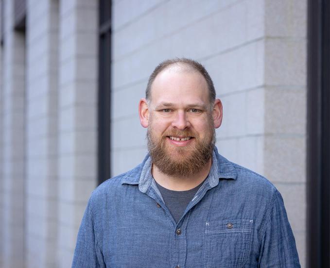 Past CU Denver student, Camden Farmer, wearing a chambray button down shirt smiling in fornt of building