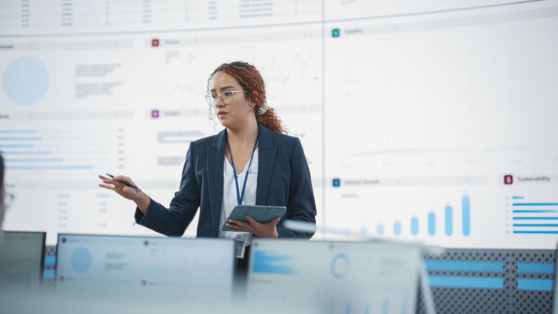 Female Data Scientist Reviewing Reports On Big Digital Screen In Monitoring Room With Employees Working Behind Desktop Computers.