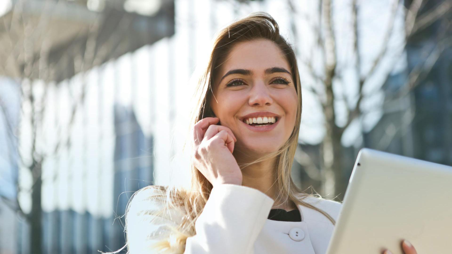 Smiling professional woman in a white coat holding a tablet and talking on the phone outdoors, with a modern cityscape in the background—representing career success in criminal justice.