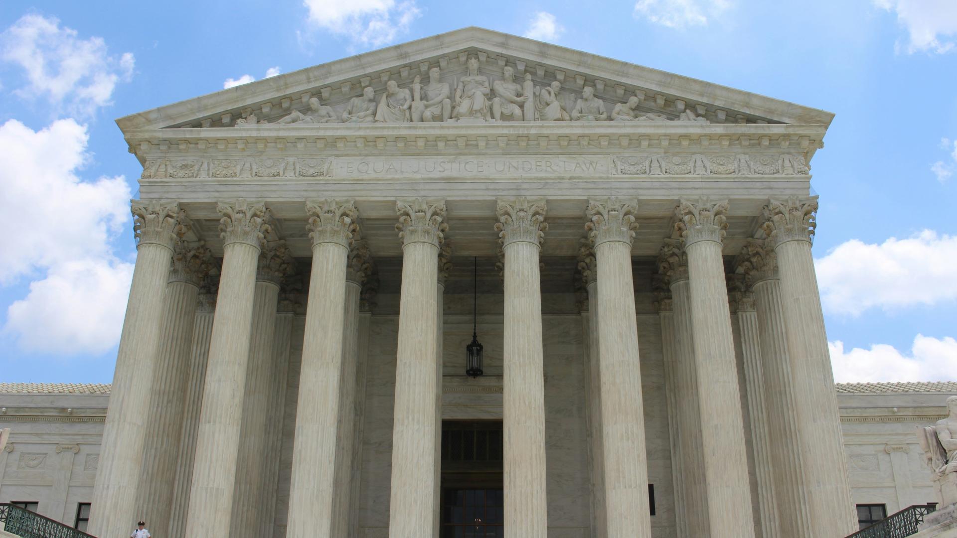 Large, white, opulent, greco-roman style, judicial building with many layered columns and row of 9 intricately carved humans at the peak with "EQUAL JUSTICE UNDER THE LAW" carved underneath. Blue sky and white clouds in the background.