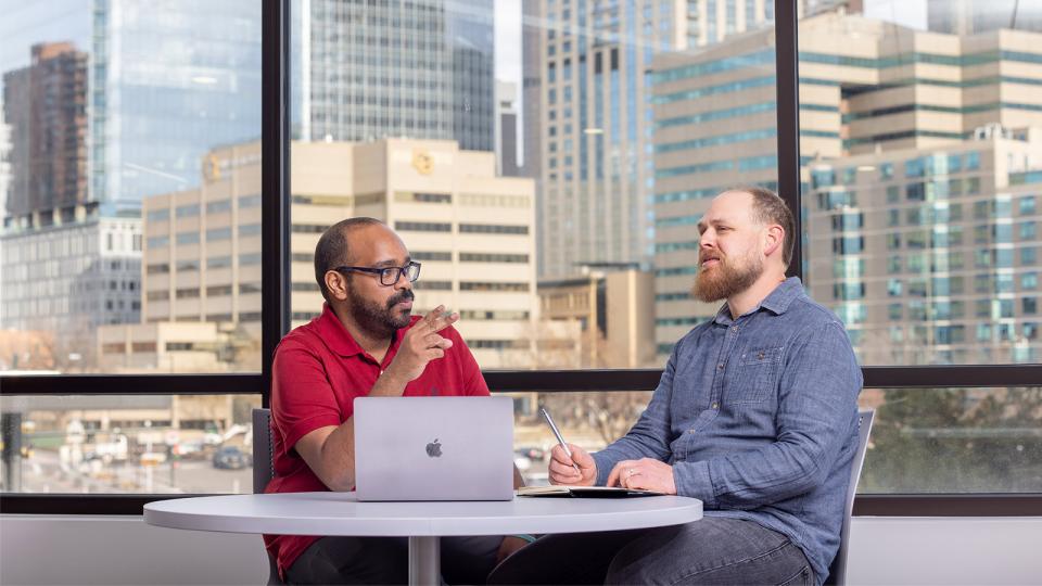 Two professionals engaged in discussion at a table with a laptop and notebook, with a cityscape visible through large windows—representing collaboration and career growth in criminal justice.