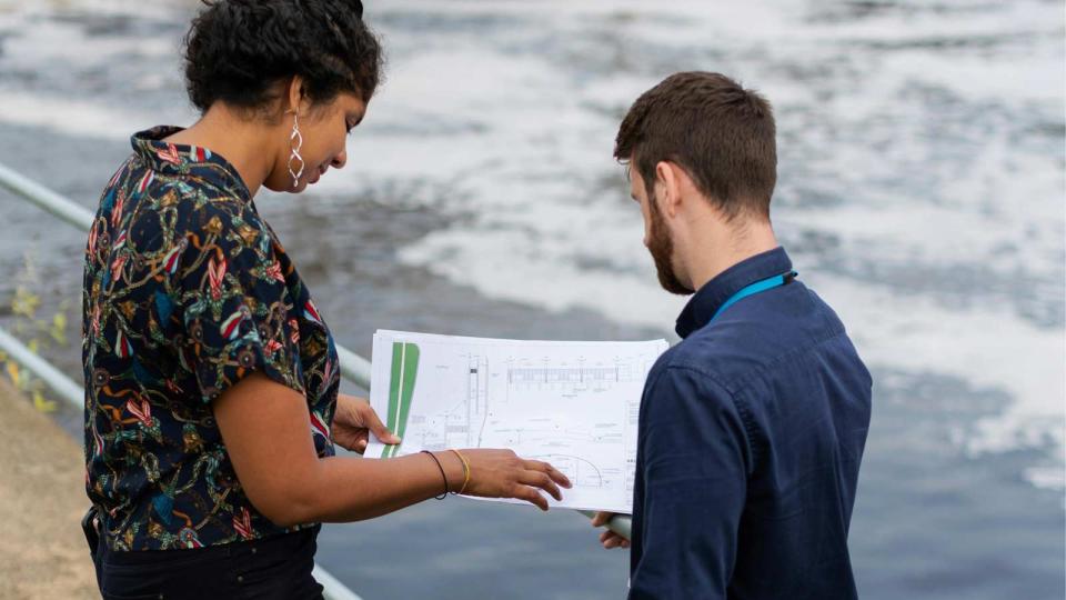 Two civil engineering professionals reviewing technical blueprints near a waterway, collaborating on infrastructure or environmental project solutions—representing innovation in engineering.