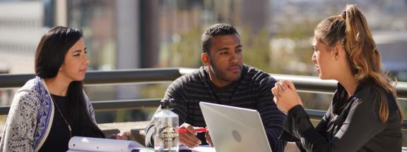 Man sitting between 2 women on a balcony at a table in front of a laptop, notebook and CU Denver branded water bottle with city buildings in the background.