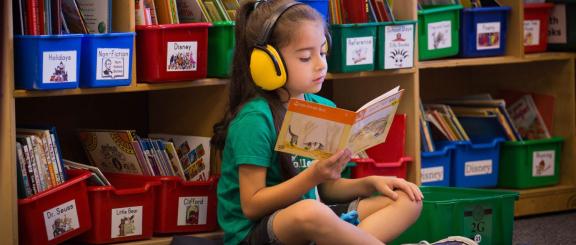 Child wearing headphones reads book in classroom.